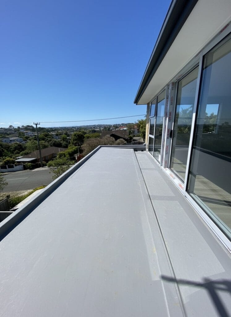 Light grey TPO waterproofed deck with modern aluminum-framed glass sliding doors overlooking suburban Auckland landscape on sunny day